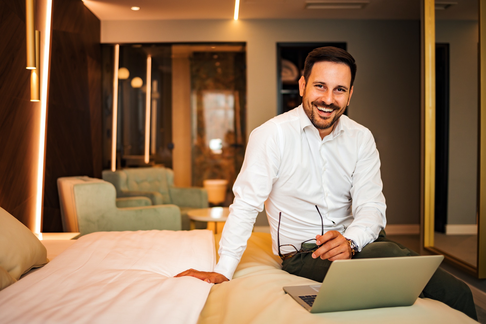 Cheerful businessman sitting on the bed of a hotel room, using laptop and smiling at camera.